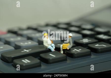 Miniature people toy figure photography. Project budget discussion. Two workers sitting above calculator. Image photo Stock Photo