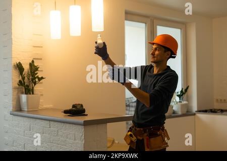 Handsome young builder in construction helmet is twisting the light bulb in. The man is looking up. Stock Photo