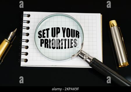 Business and finance concept. On a black surface there is a pen, a notebook and a magnifying glass. The inscription in the notebook - SET UP YOUR PRIO Stock Photo