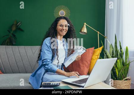 A young beautiful Latin American woman teacher of mathematics, informatics teaches online at home. He sits on the sofa with a laptop, calculator, papers, talks to students via video call, smiles. Stock Photo