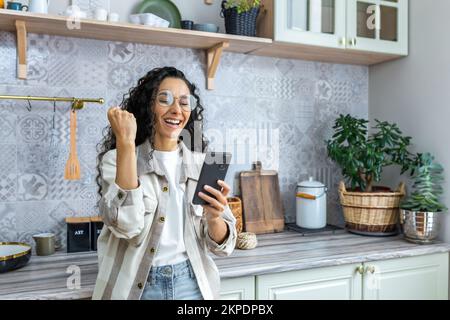 Happy beautiful young hispanic woman messing around in the kitchen at home with her phone, playing online games, winning a prize. Shows a victory gesture with his hand. Stock Photo