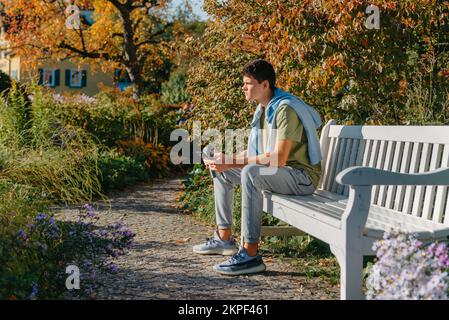 A Teenager Sits On A Bench In The Autumn Park Drinks Coffee From A Thermo Mug And Looks Into A Phone. Portrait Of Handsome Cheerful Guy Sitting On Stock Photo