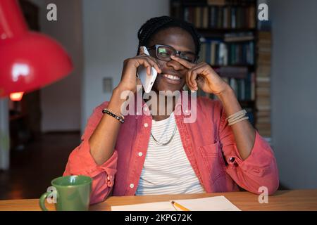 Laughing African student guy talking on smartphone, being distracted by phone while studying at home Stock Photo