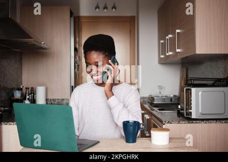 Smiling black woman talking on smartphone while using laptop Stock Photo