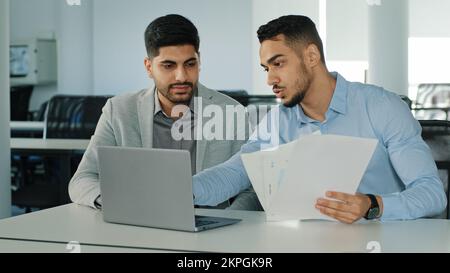 Diverse male employees Arab young colleagues discussing working together, checking emails. Two serious focused East Indian businessmen sitting in Stock Photo