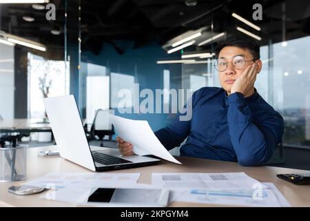 Serious thinking asian man working with documents inside office, concentrated businessman with contracts and bills reading and calculating reports, mature financier with documents. Stock Photo