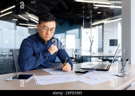 Serious thinking asian man working with documents inside office, concentrated businessman with contracts and bills reading and calculating reports, mature financier with documents. Stock Photo