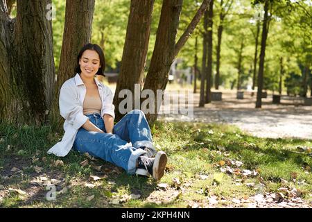 Young people. Beautiful asian girl sits near tree in park and rests, smiling and looking into distance, relaxing outdoors on fresh air in summer Stock Photo