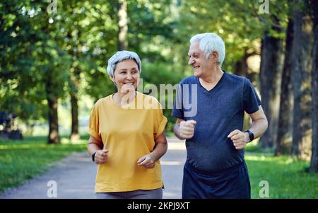 Mature smiling grey-haired fit couple jogging in the summer park. Two older joggers enjoy morning active cardio outdoors, keep healthy active lifestyl Stock Photo