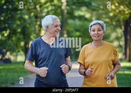 Mature fit couple jogging in the summer park. Two older joggers enjoy morning active cardio outdoors, keep healthy active lifestyle. Weightloss, sport Stock Photo