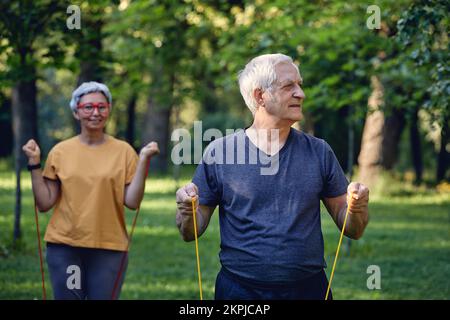 Senior 60 spouses wear sportswear doing exercises, training arms outdoor in summer park in the morning using resistance rubber bands. Healthy lifestyl Stock Photo