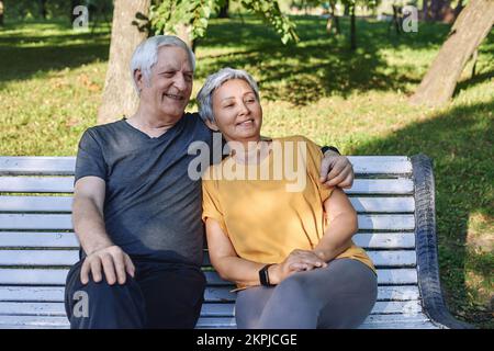 Happy multi racial elderly grey hair couple resting sit on bench in park after stroll, morning walk smiling enjoy relaxation outdoors at sunny summer Stock Photo