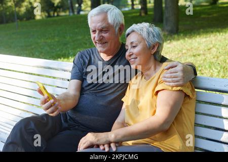 Mature spouses sit on bench in summer park with smart phone. Grey-haired couple look at cellphone screen enjoy new application outdoor use modern tech Stock Photo