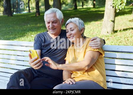 Senior spouses sit on bench in summer park with smart phone. Grey-haired couple look at cellphone screen enjoy new application outdoor use modern tech Stock Photo