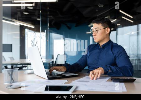 Mature Asian businessman works with documents and calculates bills uses calculator, auditor checks contracts and financial papers serious and focused. Stock Photo