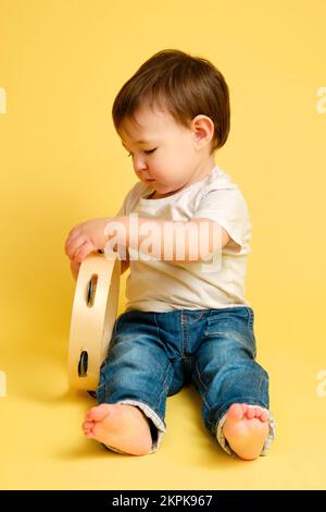 Toddler baby plays the tambourine, a child with a percussion musical instrument on a studio yellow background. Happy child musician playing hand drum. Stock Photo