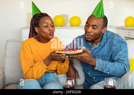 Excited african american spouses in festive hats blowing lit candles on pie, celebrating b-day together, sitting on sofa Stock Photo