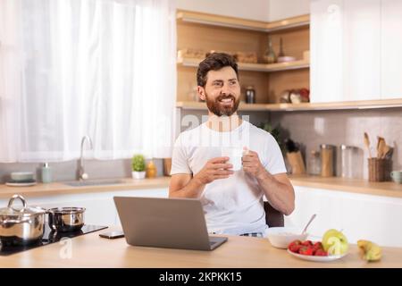 Glad adult caucasian guy with beard in white t-shirt enjoys cup of coffee, sits at table with laptop, thinks Stock Photo