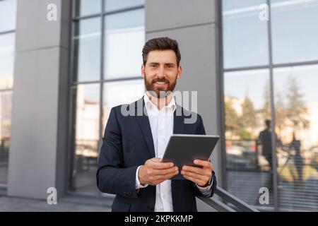 Cheerful confident attractive young european male ceo manager with beard in suit typing on tablet Stock Photo