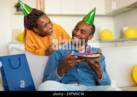 Loving black wife making surprise for her husband, happy man holding birthday pie while sitting on sofa at home Stock Photo