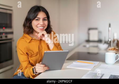 Smiling Young Arab Female Freelancer Using Digital Tablet While Sitting In Kitchen Stock Photo