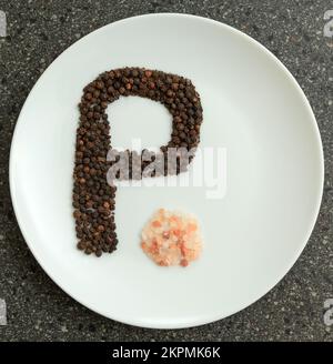Black peppercorns placed in the shape of the letter P and Pink Himalayan salt on a white plate Stock Photo