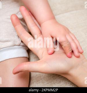 Woman mother sticks a medical band-aid on the toddler baby finger. Mom s hand with a sticky wound protection bandage and a child hand. Kid aged one ye Stock Photo