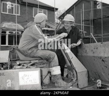 1980s, outside on a construction site, two workmen having a conversation, one, possibly a foreman wearing a hard hat, and a worker wearing a bobble hat, who is on a tip dumper, a construction vehicle that is a type of dumper truck, England, UK. Stock Photo