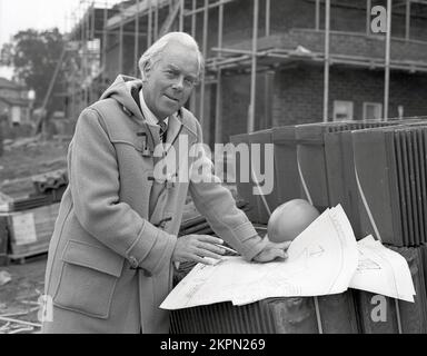 1980s, historical, a senior building contractor standing outside on a construction site of new homes, wearing a hooded duffle coat with toggle horn fastenings over a suit & tie, with technical drawings of the site laid over some roof tiles, England, UK. Stock Photo