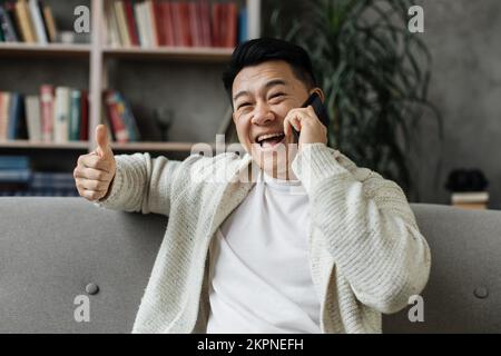 Handsome asian man in casual wear talking on smartphone while resting on comfy couch. Young male having mobile conversation during free time at home showing thumb up Stock Photo