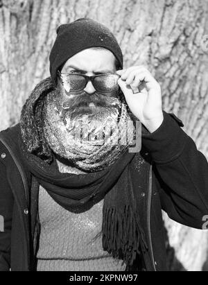 Frown bearded man, hipster, with beard and moustache in hat, scarf looks from under black sunglasses covered with white frost near tree on winter day Stock Photo