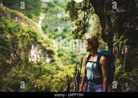 Description: An athletic woman with backpack views into the distance from the adventurous jungle path along a green overgrown canal. Levada of Caldeir Stock Photo