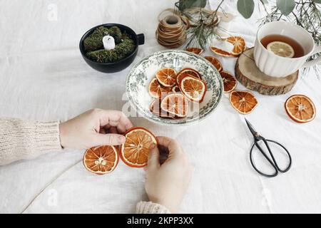 Closeup of womans hands making Christmas garland. Cup of tea, dry oranges fruit and pine, eucalyptus tree branches on white linen table cloth. Festive Stock Photo
