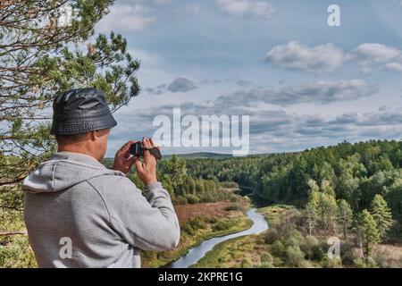 Back view of senior male hiker who is admiring landscape and filming with video camera on top of rock. Nature park Deer streams, Sverdlovsk region, Ru Stock Photo