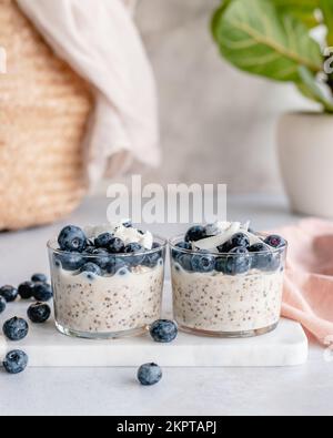 Two cups of overnight oats and chia seeds with fresh blueberries and coconut flakes on marble chopping board Stock Photo