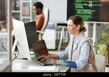 Smiling woman with bionic arm coding on computer when working on big project at research center Stock Photo