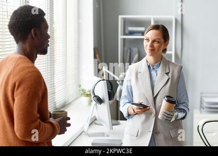 Positive businesswoman with bionic arm drinking coffee when talking to colleague Stock Photo