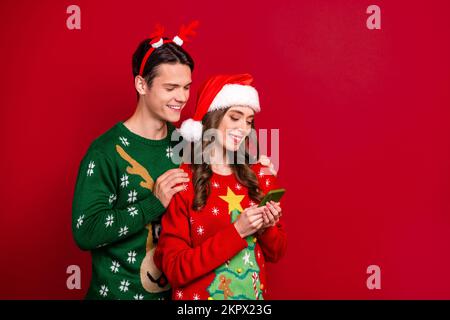 Portrait of two people embrace use telephone eshop media app santa headwear reindeer antlers isolated on red color background Stock Photo