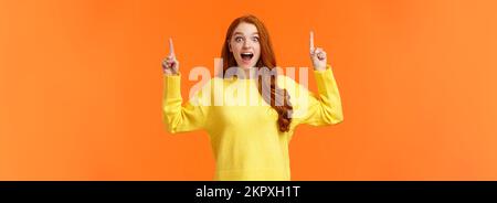 Excited girl travel abroad during winter holidays, time pack luggage. Astonished and cute redhead woman express happiness, pointing up at sale Stock Photo