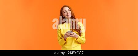 Girl searching gifts for holidays online. Carefree happy redhead female student using smartphone app, laughing joyfully, holding mobile phone Stock Photo