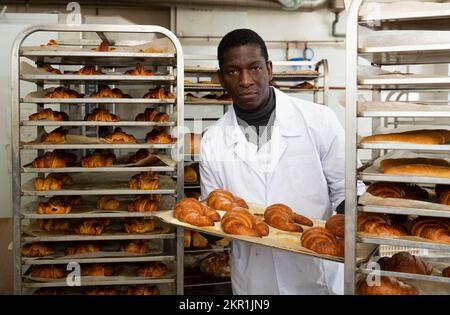 African baker arranging trays with bakery products on trolley Stock Photo