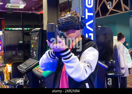 A boy playing a virtual reality video game in an arcade, holding a gun and wearing a VR headset Stock Photo