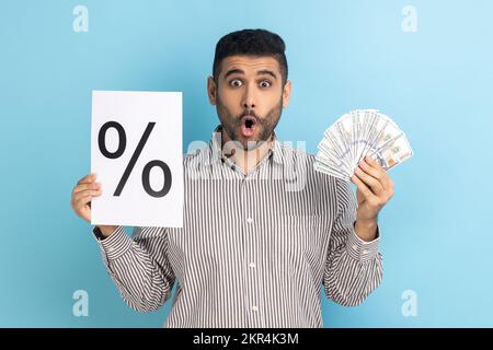 Astonished surprised businessman holding in hands percent mark on paper and dollar banknotes, looking at camera with open mouth, wearing striped shirt. Indoor studio shot isolated on blue background. Stock Photo
