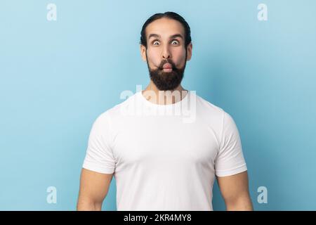 Portrait of childish man with beard wearing white T-shirt making fish face or sending kiss, having fun, kidding, pretending to be stupid. Indoor studio shot isolated on blue background. Stock Photo