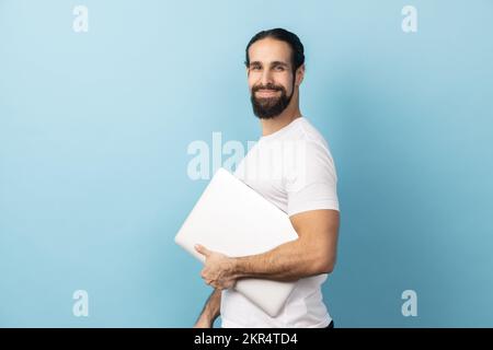 Side view of man with beard wearing white T-shirt standing, holding closed laptop or folder, looking at camera with kind look, optimistic expression. Indoor studio shot isolated on blue background. Stock Photo