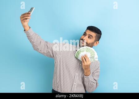 Rich businessman holding money and taking selfie by mobile phone, smiling boasting of big profit, successful online betting, wearing striped shirt. Indoor studio shot isolated on blue background. Stock Photo