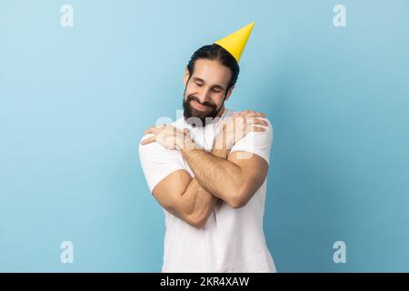Portrait of smiling handsome birthday man with beard wearing white T-shirt and party cone, celebrating holiday, hugging himself, egoist. Indoor studio shot isolated on blue background. Stock Photo