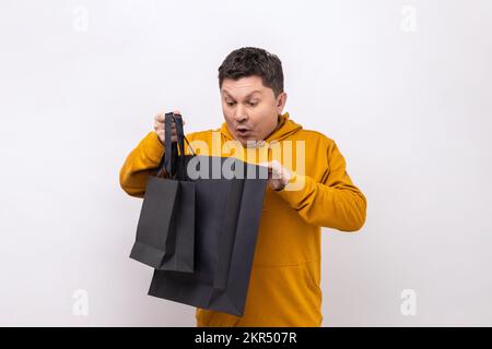 Portrait of middle aged man standing holding black shopping bags and looking inside with shocked facial expression, wearing urban style hoodie. Indoor studio shot isolated on white background. Stock Photo