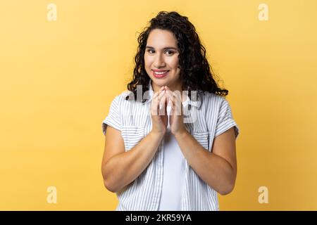 Portrait of attractive devious cunning woman with dark wavy hair clasping hands and smirking mysteriously, scheming cheats, evil prank. Indoor studio shot isolated on yellow background. Stock Photo