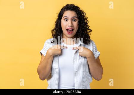 Portrait of woman with dark wavy hair pointing herself, looking with surprised expression, expressing shock, unbelievable success. Indoor studio shot isolated on yellow background. Stock Photo